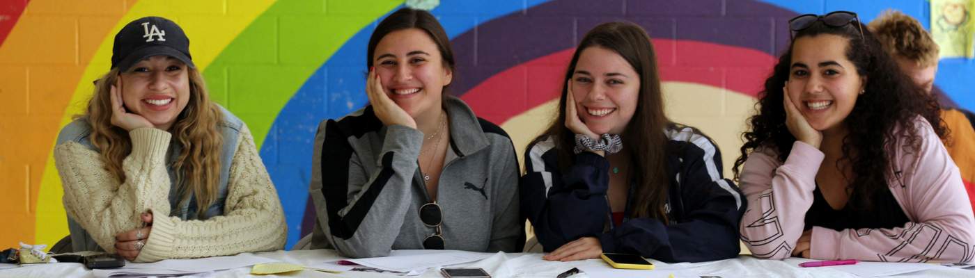 Photograph of 4 female students with rainbow painting in the background. 